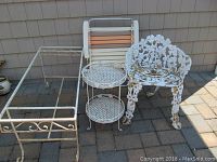 Cast iron chair, metal table frame, two-tier rack, and folding lawn chair arranged outdoors on a paved surface.