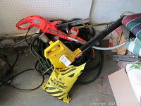 Photo showing multiple garden tools including a red and black Black and Decker electric hedge trimmer, a yellow electric leaf blower or trimmer, hoses, and a bag of Morton Safe-T-Salt in a garage.