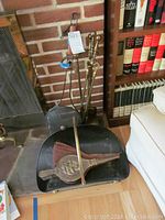 Fireplace tool set arranged with holder, brush with brass emblem, shovel, poker, and tongs near brick fireplace.