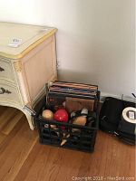 Full view of crate with vinyl albums, maracas, and tambourine on floor next to cream colored cabinet