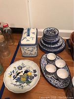 Blue-themed dishware including scalloped dish, rectangular lidded container, stacked plates and bowls, and oval tray with eggcups.