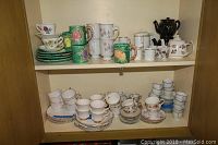 Shelf holding multiple sets of plates, various cups and mugs, and teapots with floral and fruit designs, showing the variety and quantity of china and teaware