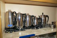Six vintage electric coffee percolators of varying sizes with black handles and stainless steel bodies on shelf.