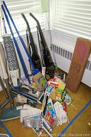 Photo showing multiple upright vacuums including two black Hoover Encore models, Electrolux Regina upright and a teal Electrolux canister vacuum in background, along with brooms and cleaning supplies on carpet near window.