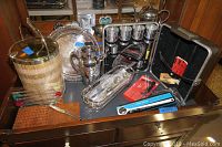 Overview of silverplate bar accessories on a wooden dresser including ice bucket, pitcher, platter, cocktail shaker, swizzle sticks, and portable bar set.