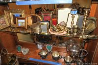 Two glass shelves with assorted silverplate and pewter items including candlesticks, photo frames, trinket boxes, teapot set, and bud vase.