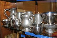 Shelf with multiple silverplated pitchers, steins, creamers, and bowls arranged in a row.