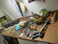 Photo of workbench with metal toolbox, hand saws, jigsaws, toilet auger, rubber mallet, files, screwdrivers, and a yellow plastic organizer tray containing small hardware components.