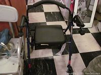 Dark blue rollator walker with black padded seat and arm rests, two front wheels, placed near a white plastic shower seat and portable commode in background.