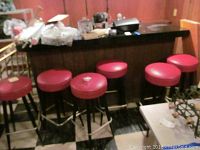Overview photo showing six red vinyl cushioned round seat bar stools with black metal legs and silver footrests, arranged in a row at a wooden bar counter.
