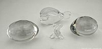 Four clear glass paperweights: two oval fish-etched, whale form, whimsical figurine