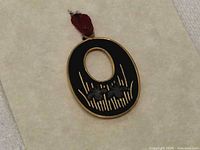 Front view of oval black iron pendant with gold and silver landscape motif and red loop, on display card
