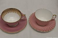 Two matte pink Aynsley teacup-and-saucer sets with gold trim displayed side by side