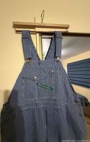 Front upper section of blue/white hickory-stripe bib overalls on hanger showing bib pocket and Key logo tag