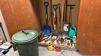 Overview of trash bin, long-handle tools, watering cans and assorted hand tools