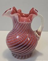 Oblique view of cranberry glass pitcher showing spiral stripes, ruffled rim, clear handle, footed base