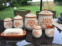 Full view of set on black table showing all seven pieces outdoors, five lidded jars, labeled cream jars, butter dish, and creamer with orange floral and bird designs