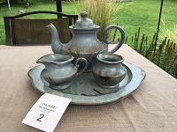 Full tea service set on table showing teapot, creamer, sugar dish on round serving tray with outdoor background.