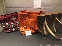 Three handbags side by side on a table: a burgundy handbag with zebra print and studded decorative cross, an orange crocodile-patterned handbag, and a beige handbag with G logos and light colored handles.