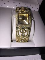 Front view of the Isaac Mizrahi ladies fashion watch with gold-tone metal and clear rhinestones, displayed on white cushion inside box.