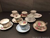 Overview of seven china tea cups and saucers of varied floral designs and gold accents on a dark surface.