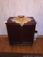 Front and top view of the storage cabinet with two doors closed, topped with a lace cloth and small bowl for scale.