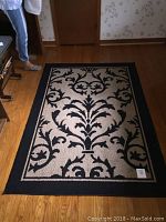 Full view of the rectangular area rug showing the black cartouche pattern on a tan background with black border and rubber backing.