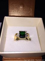 Gold ring with green rectangular stone and clear accent stones in a cream-colored ring box.