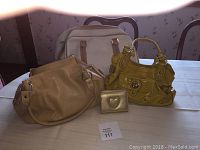 Photo showing three handbags and one small change purse arranged on table, includes beige purse, off-white overnight bag, yellow purse, and gold-tone change purse.