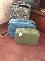 Full view of three suitcases arranged on carpeted floor near a wooden cabinet and television. Visible are floral pattern rolling suitcase, blue soft suitcase, and small green hard case suitcase.