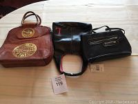 Lot of three bags on white table including reddish-brown Pink One leather bag with embroidered designs, black tote bag with 'T' on front, and black shoulder bag with silver zippers.