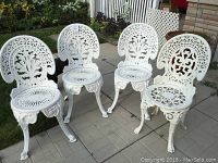 Four white cast metal ornate chairs placed on a patio, showing detailed backrests and seats with floral and scrollwork designs.