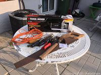 Overview of all items on white metal table including boxed Black & Decker sander polisher, orange extension cord, multiple hand tools with saw, pliers, and screwdrivers.