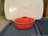 Front view of orange Le Creuset oval Dutch oven with lid, placed on blue surface against white background.