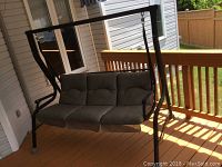 Front view of the black metal framed patio swing with grey cushion and canopy on a wooden deck balcony.