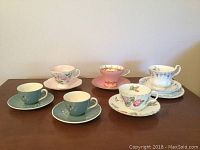 Full set of five tea cups and saucers with various colors and floral designs, displayed on wood surface.
