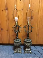 Pair of ornate table lamps shown side by side against wooden paneling. Metal body with decorative detailing, different lampshades missing, different finials on each.