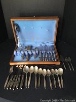 Wooden box containing 1847 Rogers Bros IS Heritage flatware pieces arranged inside with some additional flatware and utensils laid out on table
