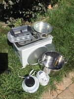 Wide view of entire lot on grass showing pot with wooden paddle, baking pans arranged on a white box, mixing bowls stacked on top, analog kitchen scale and mini electric food chopper placed in front.