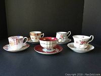 Front view showing five tea cup and saucer sets arranged with varied patterns and colors, all with floral designs and gold trim