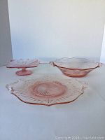 Front view of three pink glassware items: large serving tray, oval bowl, and pedestal dish with fluted edge.