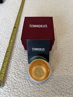 Tea bowl with gold interior beside burgundy Tenmokus box and booklet; tape measure shows size