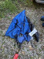 Collapsed blue tent, black sections, poles, chair frame visible on gravel