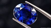 Front view of oval blue sapphire held in tweezers showing faceted crown