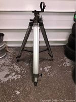 Full view of tripod standing upright showing aluminum central column and metal legs on concrete floor.