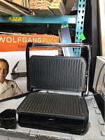 Closed and open views of the Wolfgang Puck panini maker showing ribbed removable plates and stainless steel handle, with original box and packaging in the background.