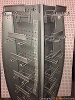 Front angle showing grey metal four sided display rack with multiple pegboard hooks on each panel against a perforated wall background.