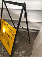 Black metal A-frame sidewalk sign holder with a yellow sign displaying "MaxSold Auction Pick-Up" text.