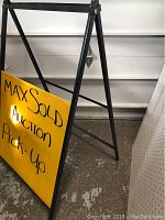 Close-up photo focusing on the lower half of the black metal A-frame sign holder and yellow sign panel with handwritten message.