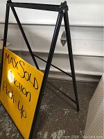 Side view photo showing black metal A-frame frame and yellow sign panel with 'MaxSold Auction Pick-Up' handwritten.